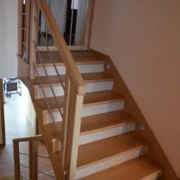 Jasne, dębowe schody z metalowymi prętami balustrady i białymi podstopnicami, oświetlone małymi lampkami w ścianie, widziane z dołu pod kątem.