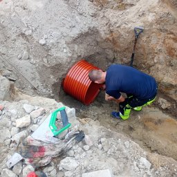 NOWAMONT Mateusz Witek - Pracownik w wykopie łączy pomarańczową rurę karbowaną, obok leży łopata i piła ręczna.