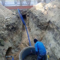 NOWAMONT Mateusz Witek - Głęboki wykop z betonowym kręgiem, wewnątrz którego hydraulik łączy niebieskie rury wodociągowe z zaworami, widoczne połączenia rur na zewnątrz wykopu.