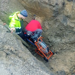 NOWAMONT Mateusz Witek - Dwóch pracowników w wykopie, jeden w neonowej kurtce przy drabinie, drugi pochylony nad pomarańczową maszyną do przeciągania kabli, widok z góry.