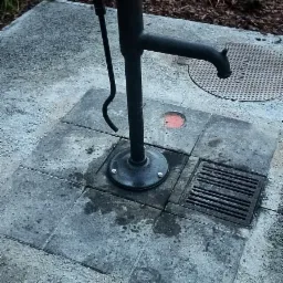 Żeliwna pompa wodna z dźwignią na brukowanym placu, obok kratka ściekowa i właz kanalizacyjny, w tle ściółka z kory.