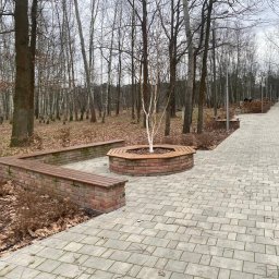 NOWAMONT Mateusz Witek - Parkowa alejka wyłożona szarą kostką brukową, z ławkami z cegły i drewna, otaczającymi drzewo z białą korą, w tle las bezlistny.