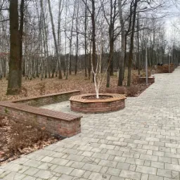 Parkowa alejka wyłożona szarą kostką brukową, z ławkami z cegły i drewna, otaczającymi drzewo z białą korą, w tle las bezlistny.