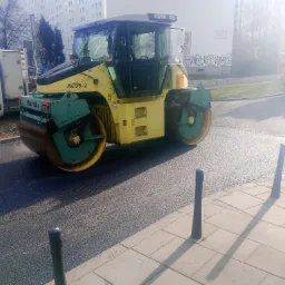 Żółto-zielony walec drogowy Ammann AV95-2 ubija świeżo wylaną nawierzchnię asfaltową na tle bloku mieszkalnego i zaparkowanych pojazdów.