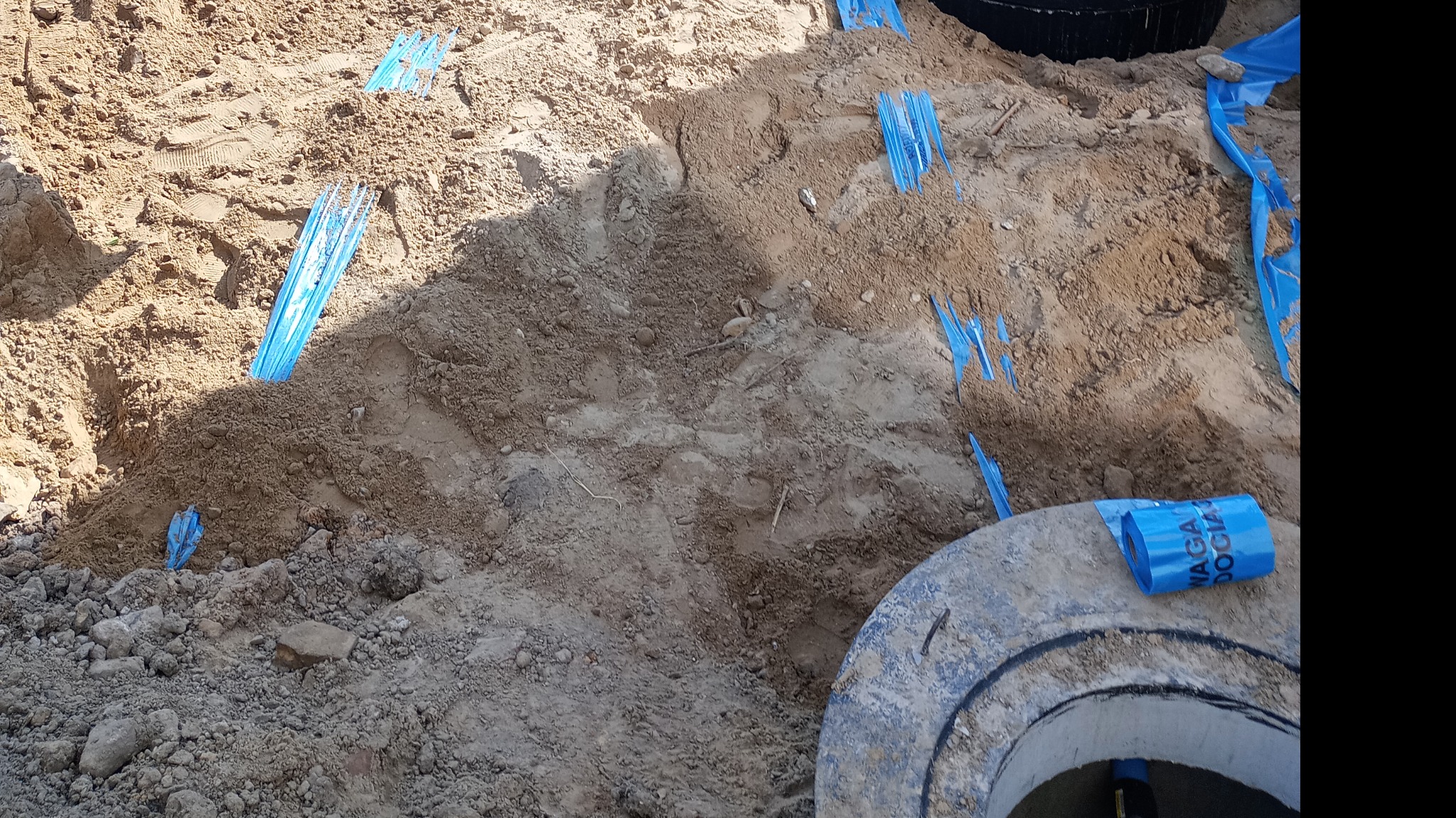 Wykonany wykop w ziemi z widocznymi niebieskimi taśmami oznaczającymi przebieg instalacji, fragment betonowego kręgu studziennego z rolką niebieskiej folii leżącą na nim.