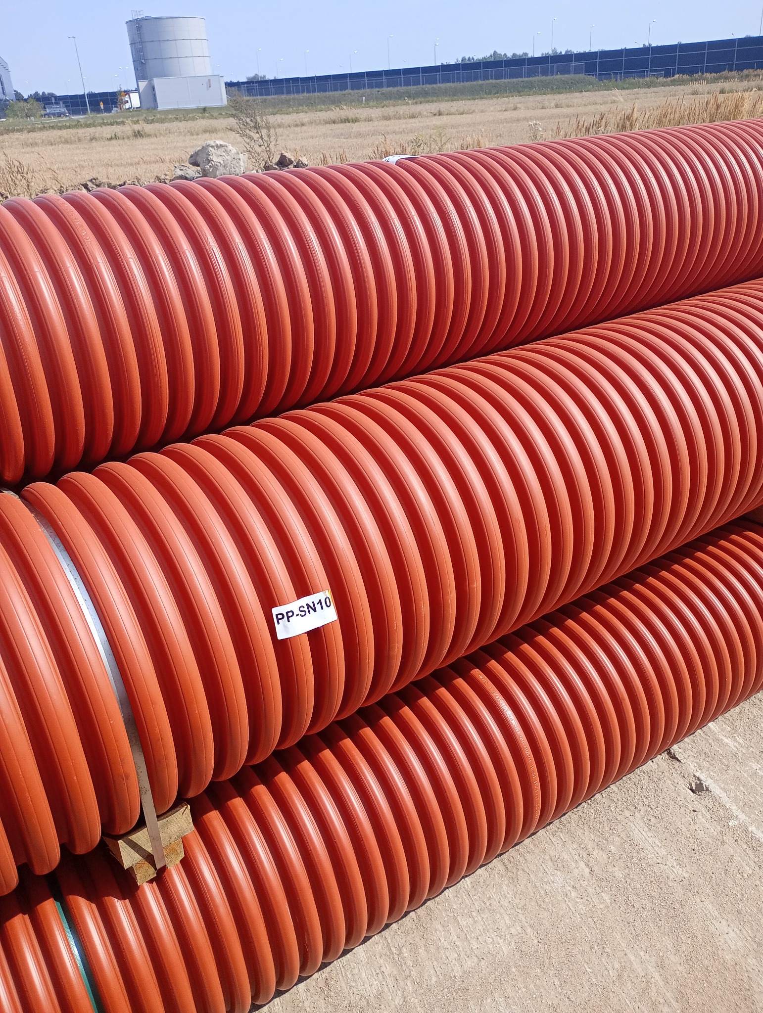 Pomarańczowe, karbowane rury drenarskie PP-SN10 ułożone jedna na drugiej na drewnianych podkładkach, widoczne w magazynie na zewnątrz.