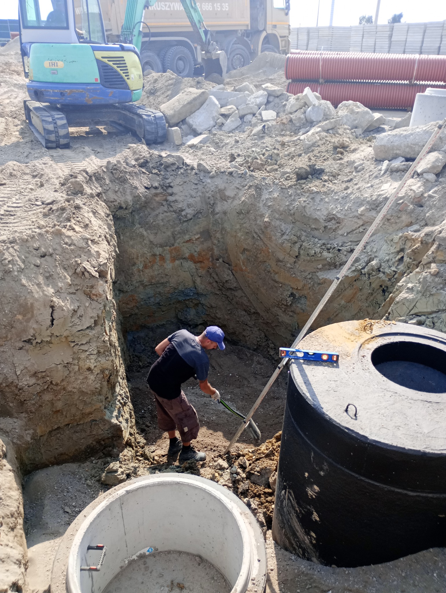 Pracownik z łopatą w wykopie pod zbiornik szamba, w tle mini koparka i rury kanalizacyjne.
