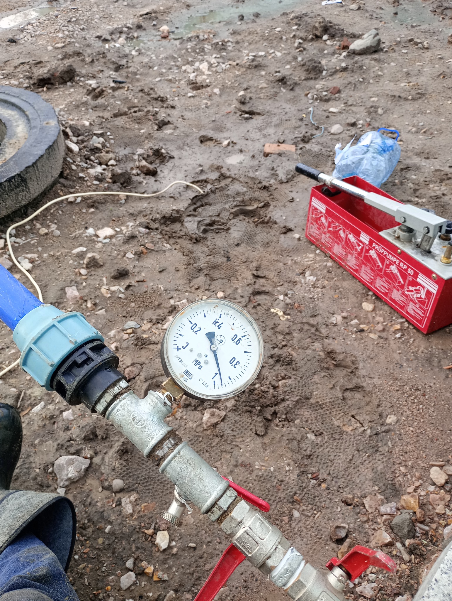 Próba ciśnieniowa rur wodociągowych na placu budowy, manometr wskazuje ciśnienie, widoczna pompa ciśnieniowa i fragment niebieskiej rury, teren wokół błotnisty.