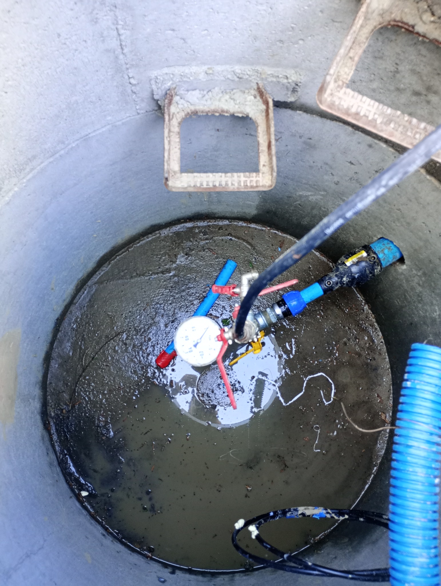 Wnętrze betonowej studni z wodą, widoczny manometr, zawory z czerwoną rączką i niebieski karbowany wąż, ujęcie z góry.