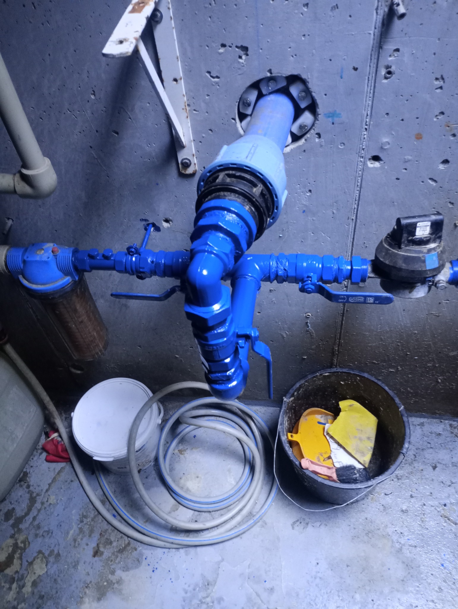 Komplet nowych, pomalowanych na niebiesko rur wodociągowych z zaworami i licznikiem, zamontowanych przy szarej betonowej ścianie, z widocznym otworem w ścianie, obok leżą zwinięte węże i wiadro...