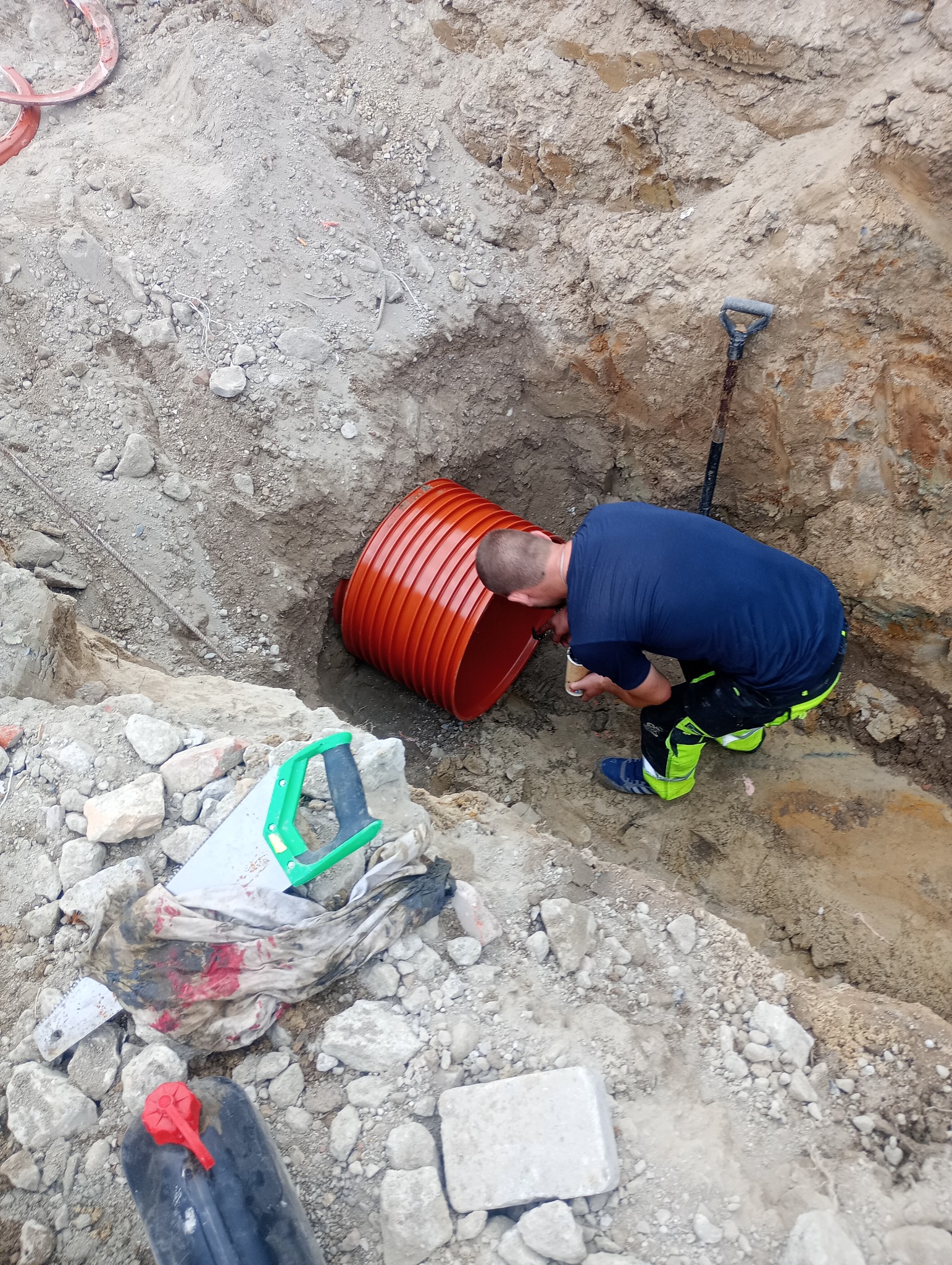 Pracownik w wykopie łączy pomarańczową rurę karbowaną, obok leży łopata i piła ręczna.