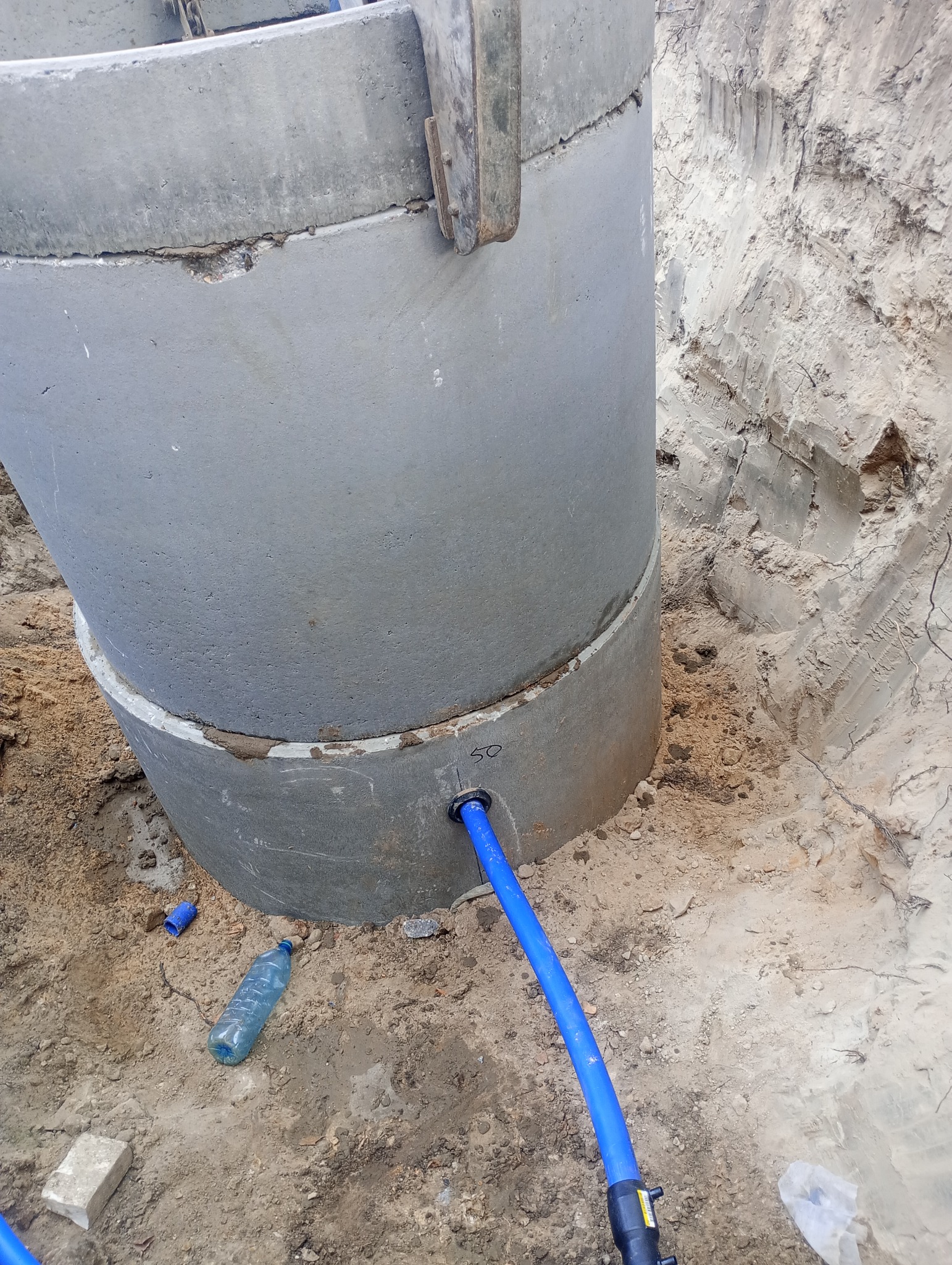 Betonowy krąg studzienny z podłączonym niebieskim przewodem w wykopie, widoczna butelka wody i numer '50' napisany na kręgu.