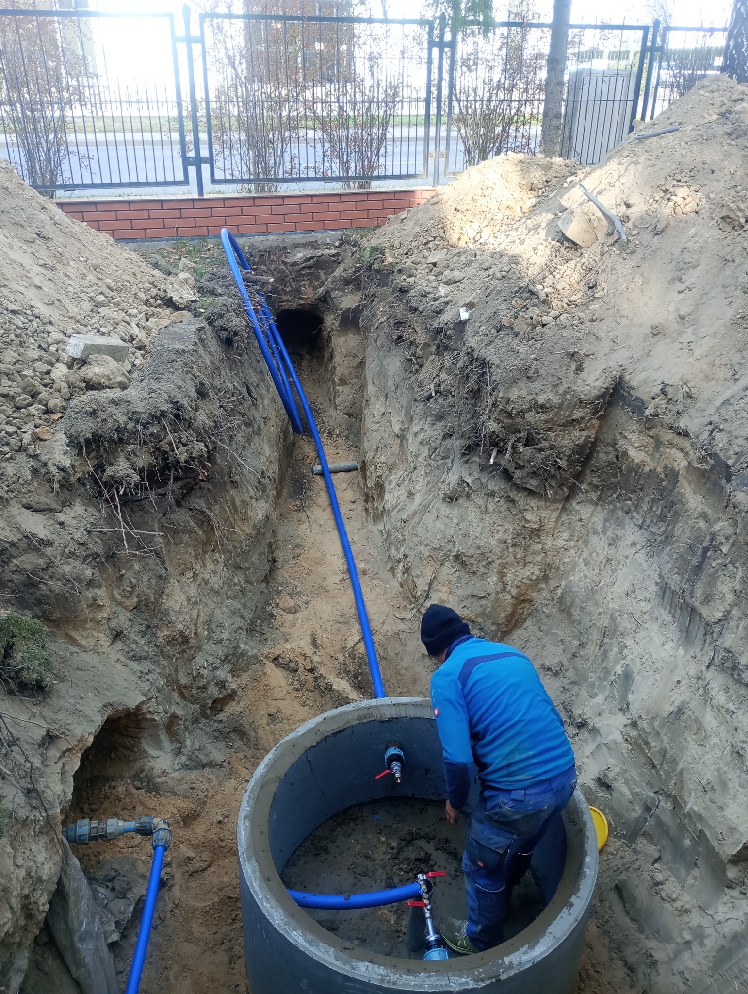 Głęboki wykop z betonowym kręgiem, wewnątrz którego hydraulik łączy niebieskie rury wodociągowe z zaworami, widoczne połączenia rur na zewnątrz wykopu.