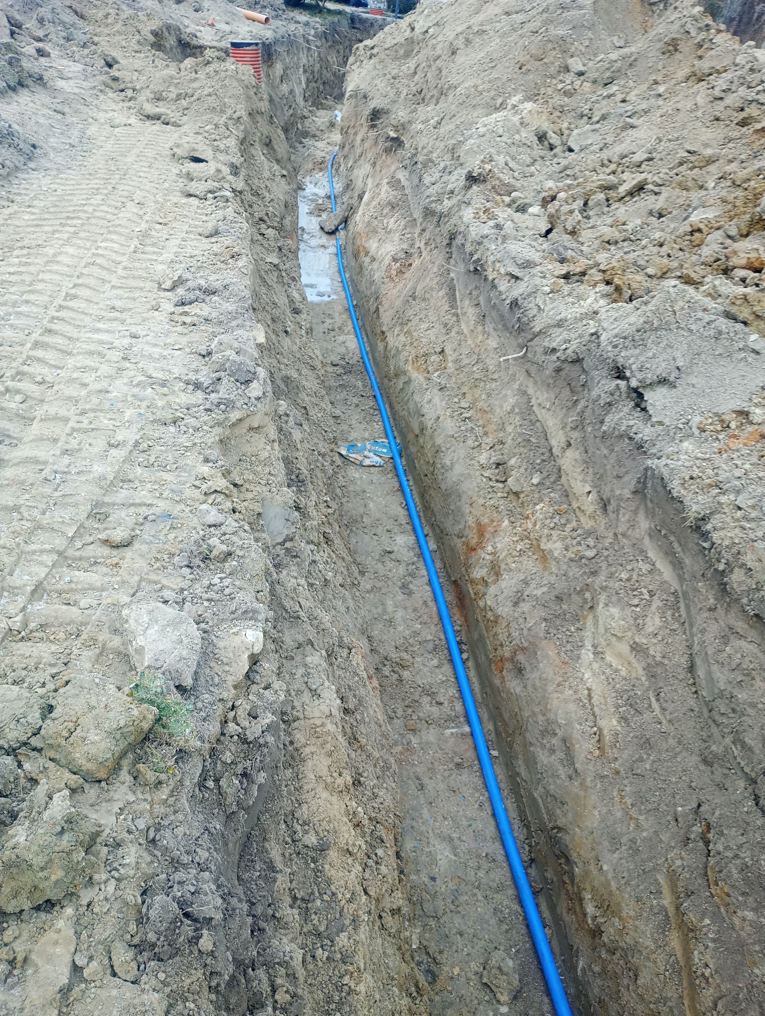 Wykopany wąski rów z ułożoną niebieską rurą wodociągową na dnie, widoczne ślady opon na ziemi obok rowu.