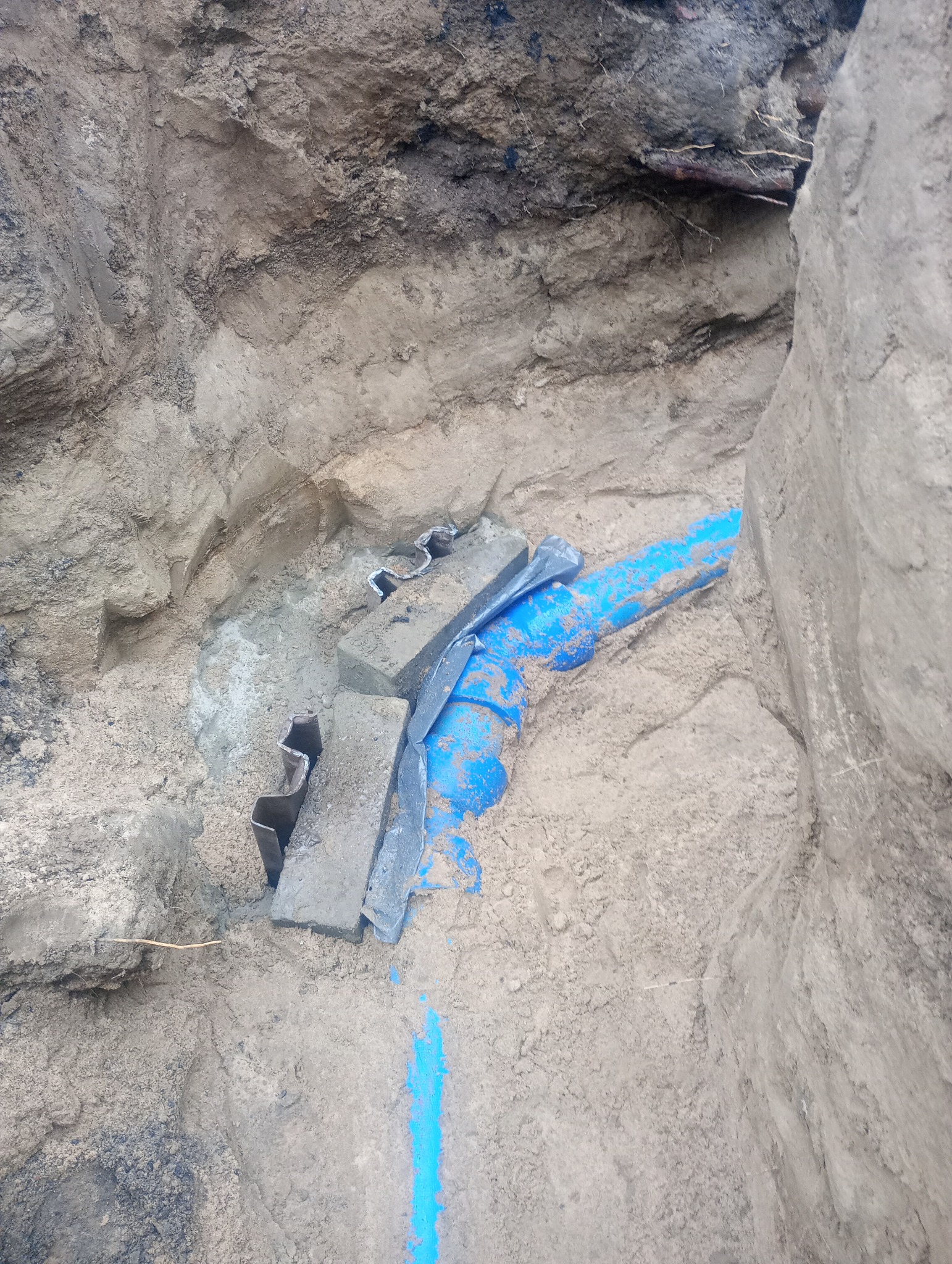 Wykop w ziemi z widoczną niebieską rurą, zabezpieczoną betonowymi blokami i metalowymi elementami, wskazującymi na etap prac instalacyjnych.