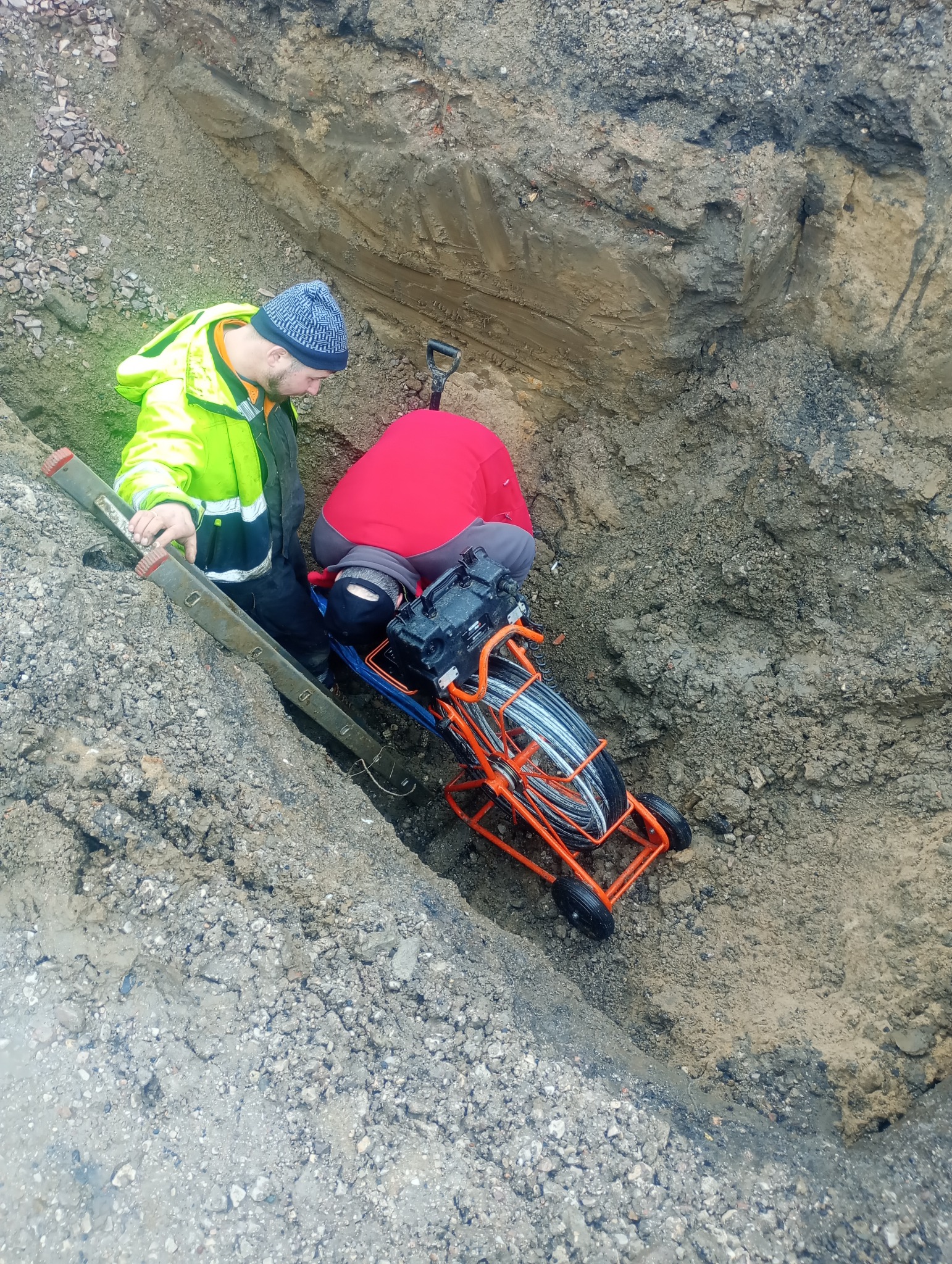 Dwóch pracowników w wykopie, jeden w neonowej kurtce przy drabinie, drugi pochylony nad pomarańczową maszyną do przeciągania kabli, widok z góry.