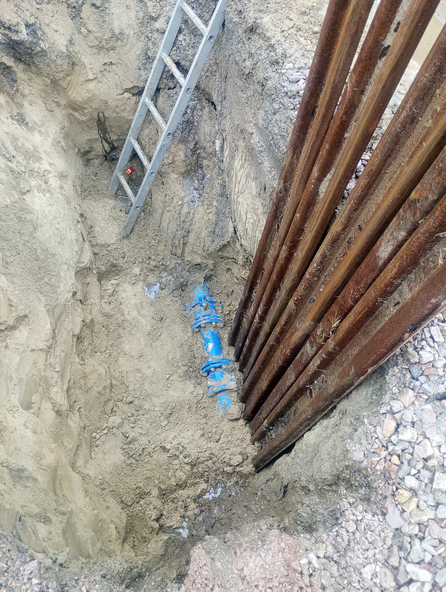 Głęboki wykop w ziemi zabezpieczony metalowymi szynami, z widocznym niebieskim przyłączem wodociągowym i aluminiową drabiną w tle.