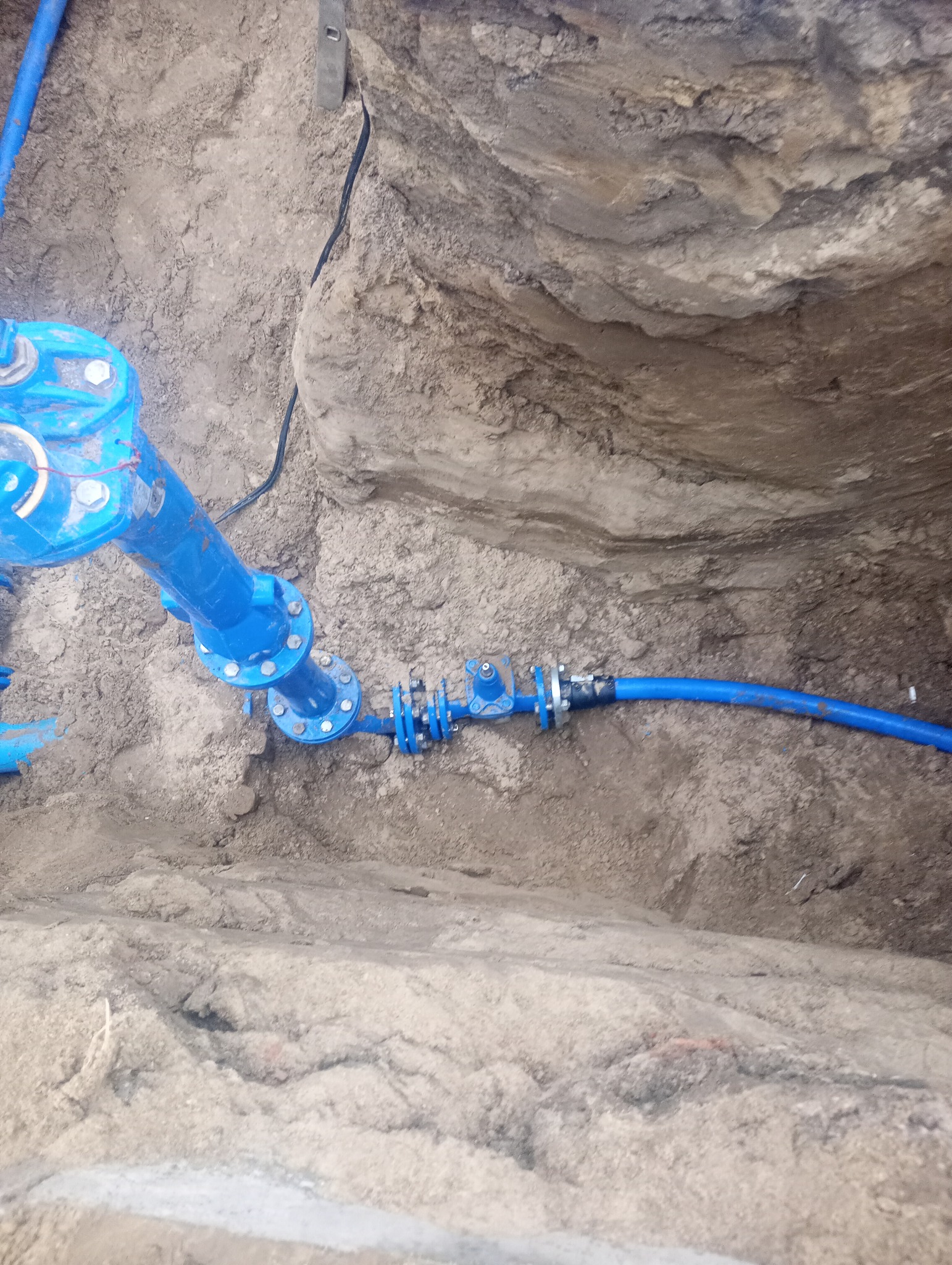 Niebieskie rury wodociągowe połączone w wykopie ziemnym, widoczne ślady świeżego piasku i poziomica przy krawędzi wykopu.