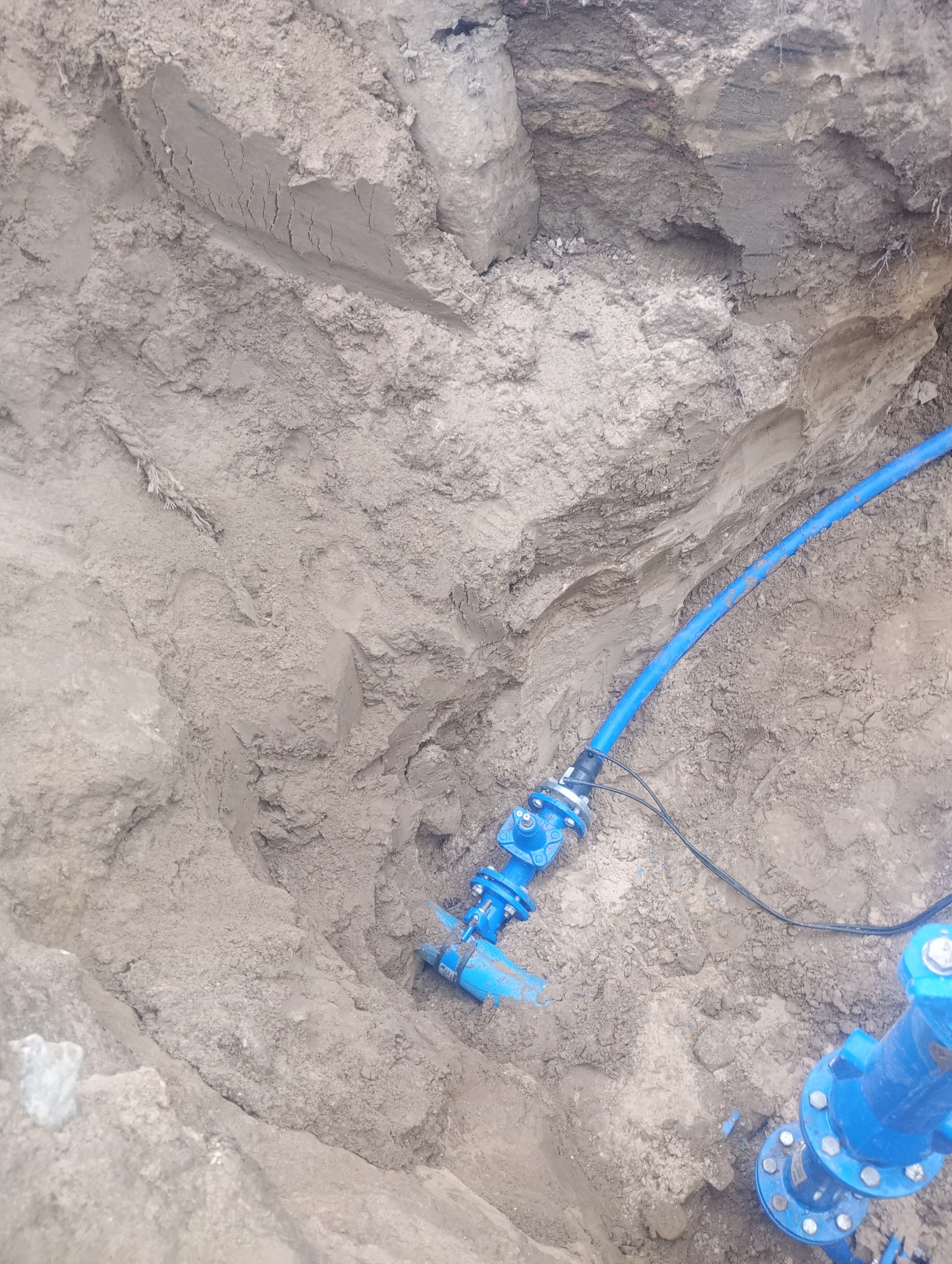 Niebieska rura wodociągowa z zaworem w wykopie ziemnym, widoczne ślady świeżych prac ziemnych.