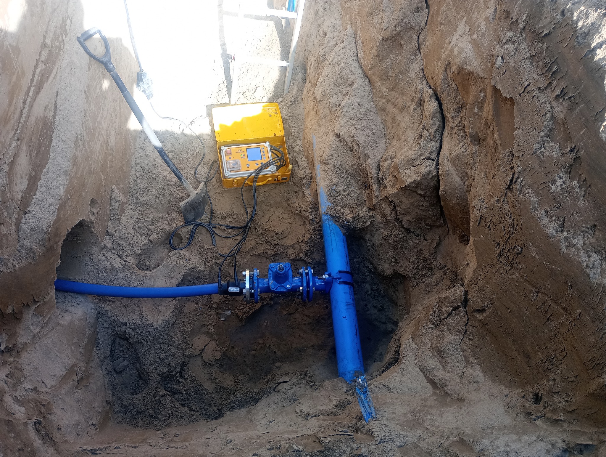 Głęboki wykop z ułożonym niebieskim rurociągiem z zaworem, żółta skrzynka z urządzeniem pomiarowym na górze, łopata oparta o ścianę wykopu.