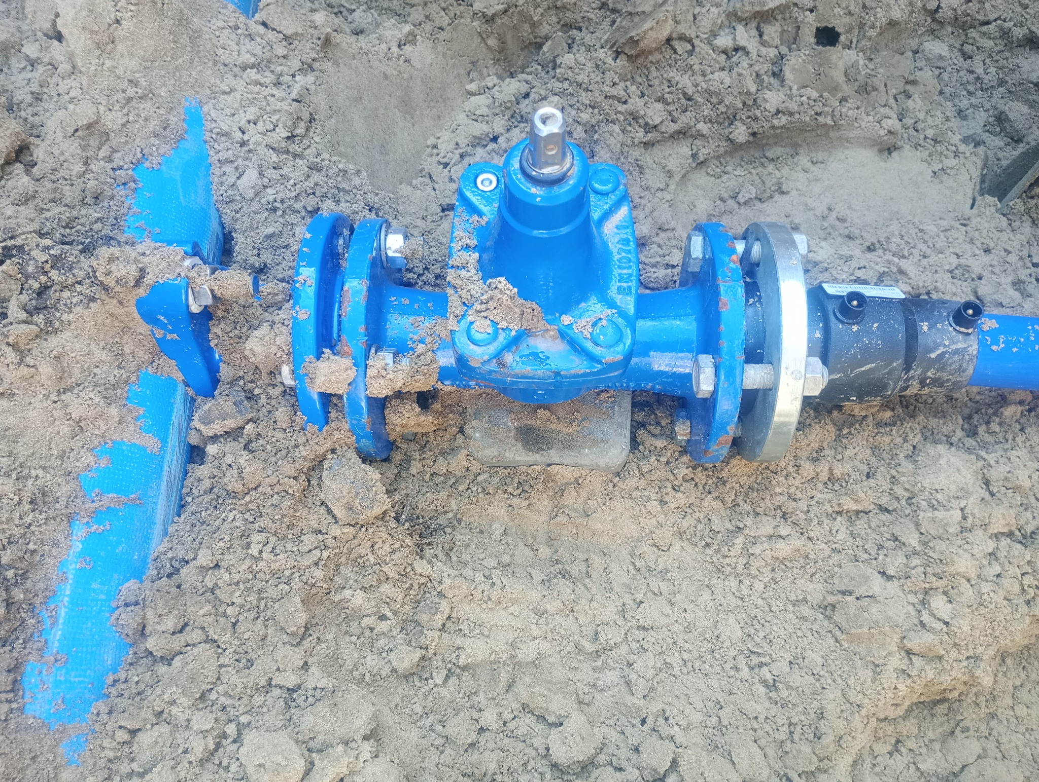 Niebieska rura wodociągowa z zaworem, częściowo zakopana w piasku podczas instalacji przyłącza wodno-kanalizacyjnego.