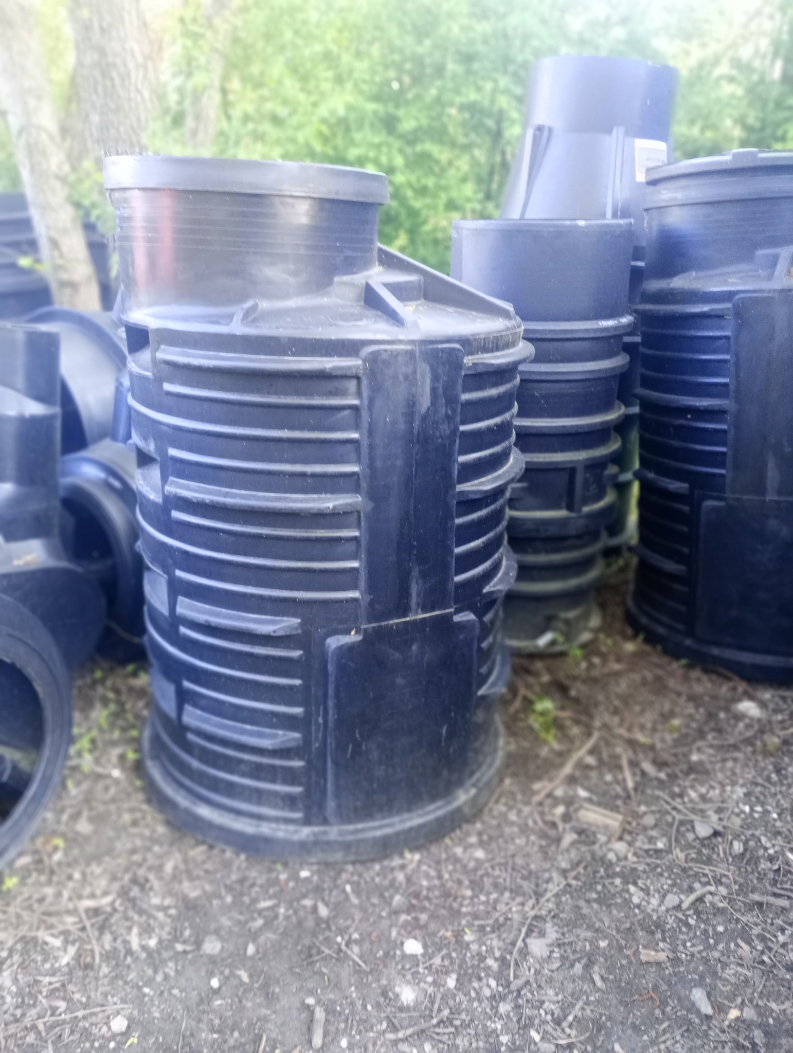 Czarne, plastikowe studzienki kanalizacyjne składowane na ziemi, z widocznym drzewem w tle.