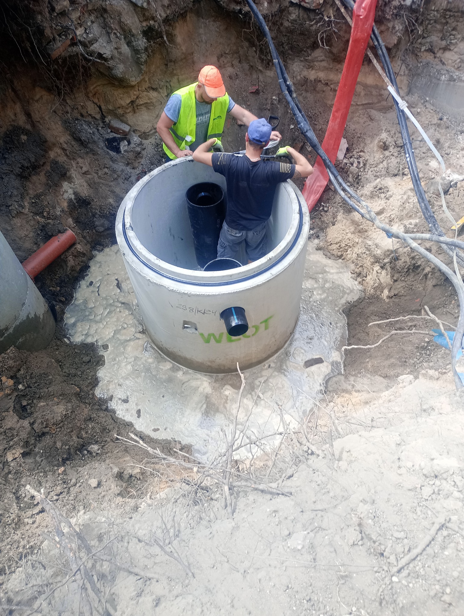 Montaż betonowego kręgu studziennego w wykopie w Piasecznie, dwóch pracowników w odblaskowych kamizelkach przy pracy, widoczne kable i rury.