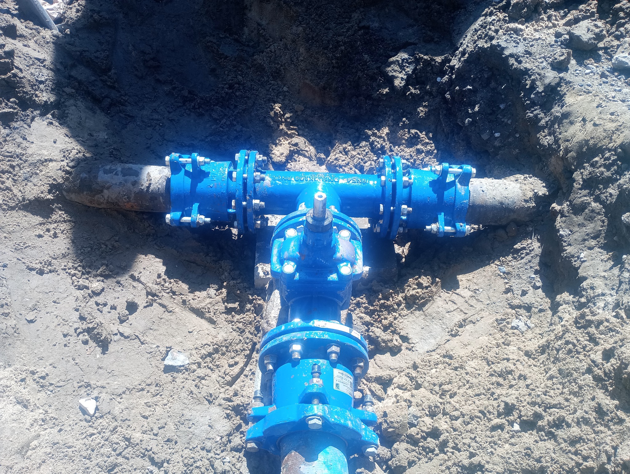 Niebieski zawór wodny z przyłączami śrubowymi, zakopany w ziemi, widoczny w wykopie.