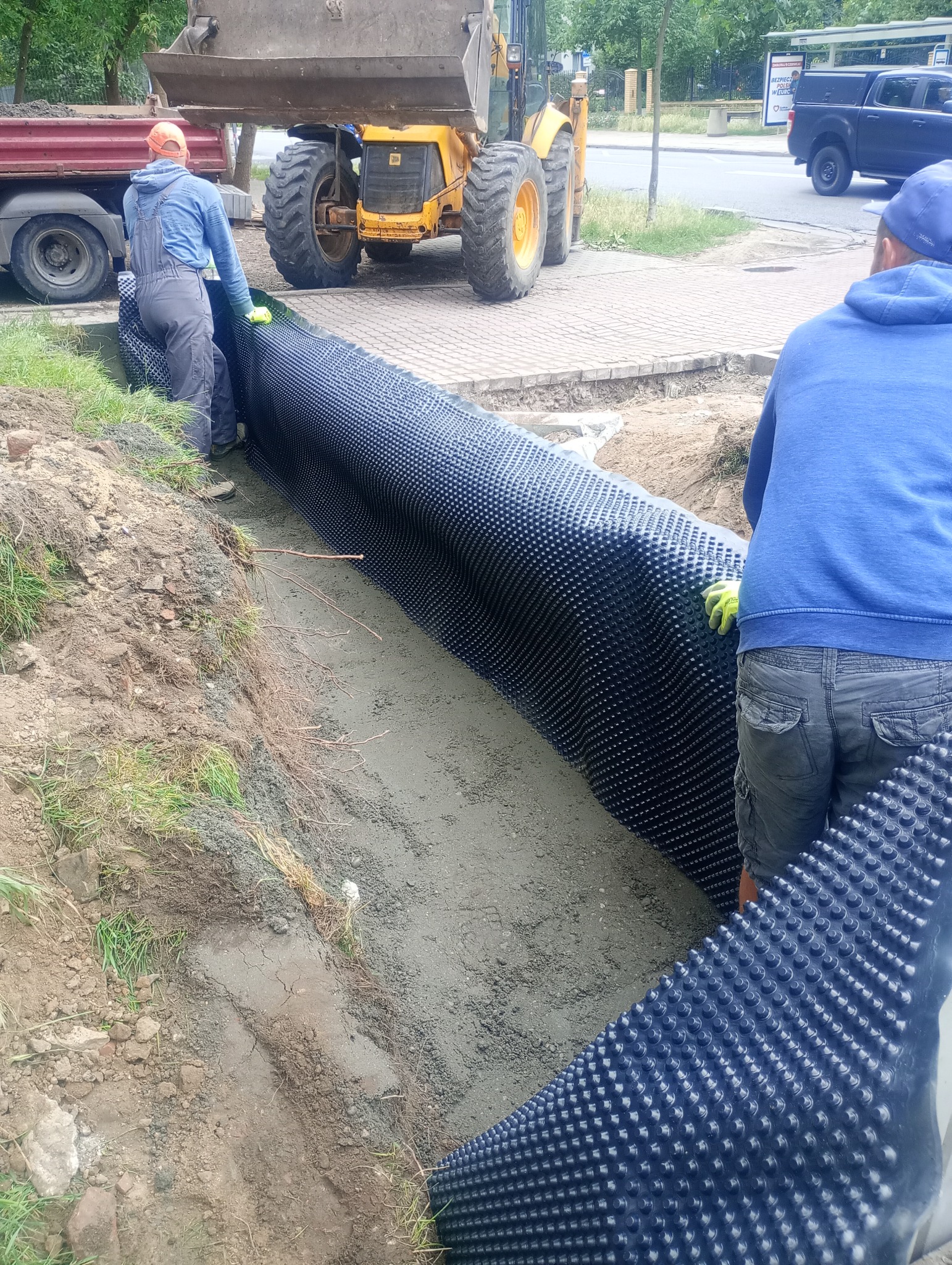 Pracownicy w żółtych rękawicach układają czarną folię kubełkową w wykopie, w tle żółta koparko-ładowarka JCB i ciężarówka na tle chodnika i zieleni miejskiej.