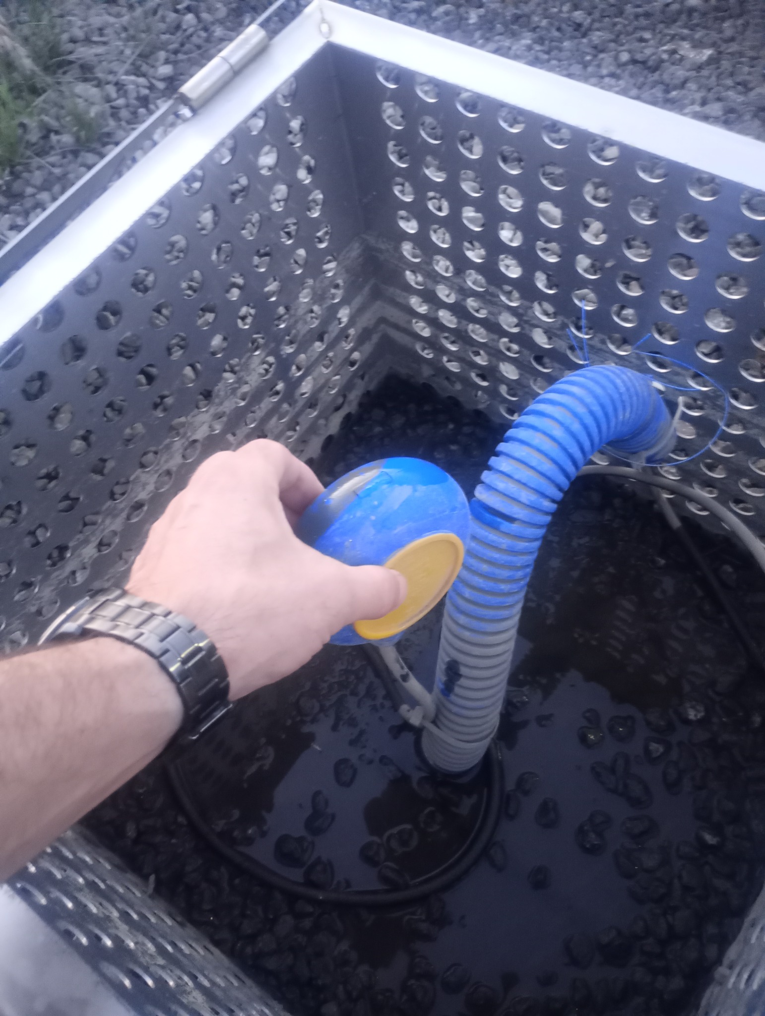 Studzienka drenażowa z perforowanymi ścianami, wypełniona żwirem, z niebieskim pływakiem i karbowanym wężem; widoczna ręka w zegarku.