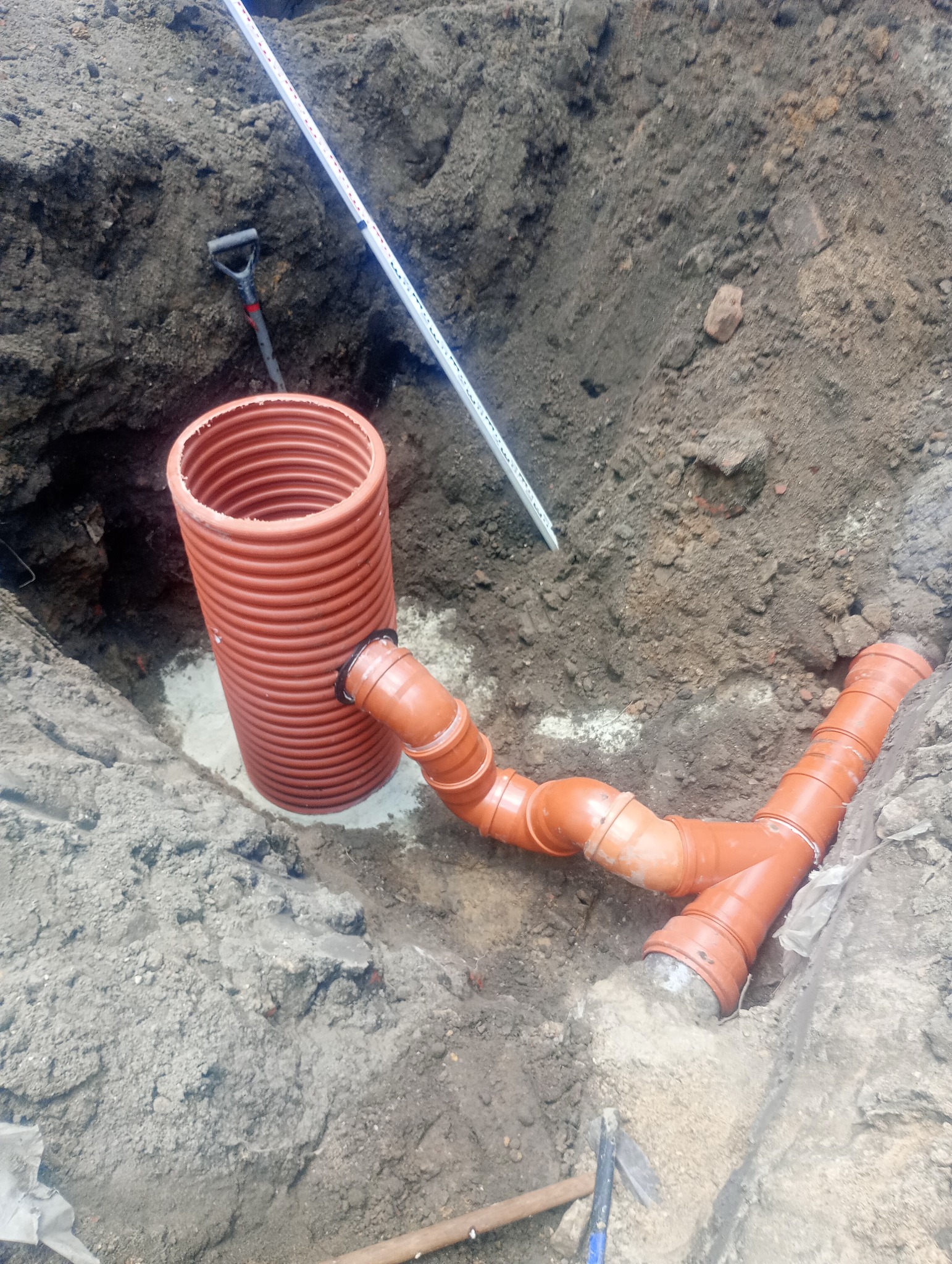 Pomarańczowe rury kanalizacyjne z trójnikiem, częściowo zasypane w wykopie, z widoczną poziomicą i szpadlem w tle.