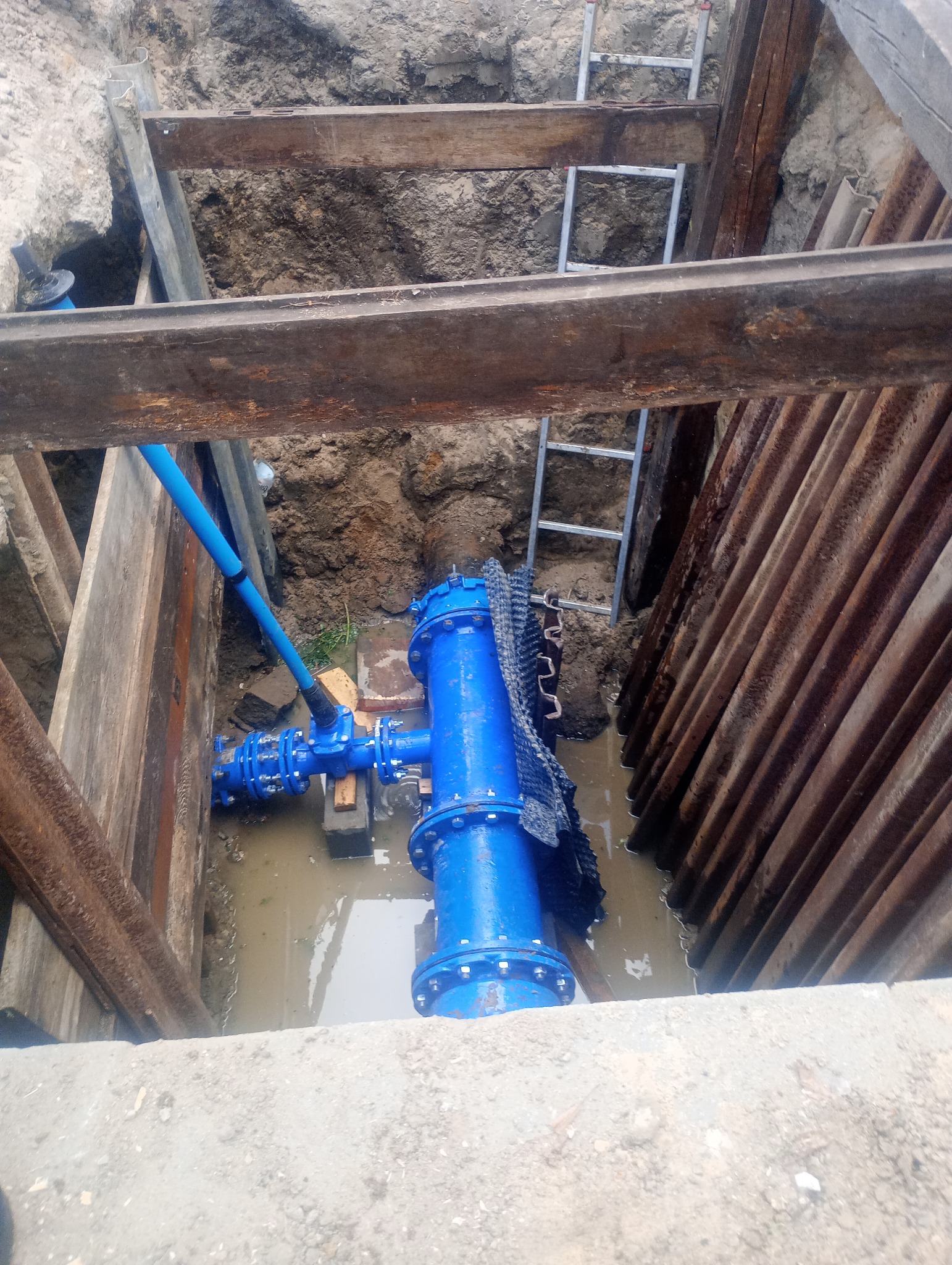 Niebieska armatura wodociągowa w wykopie zabezpieczonym drewnianą konstrukcją i stalowymi szynami, częściowo zalana wodą, widoczna drabina.