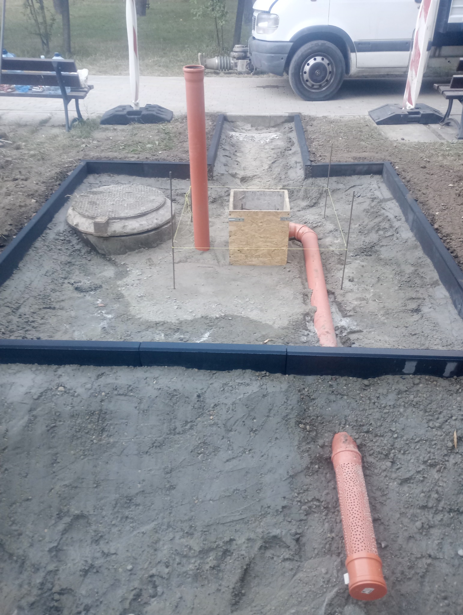 Wykonany wykop z widocznymi elementami instalacji: pionowa rura kanalizacyjna, skrzynka drewniana, rura drenarska z otworami, czarna obudowa z kostki oraz studzienka kanalizacyjna. W tle...