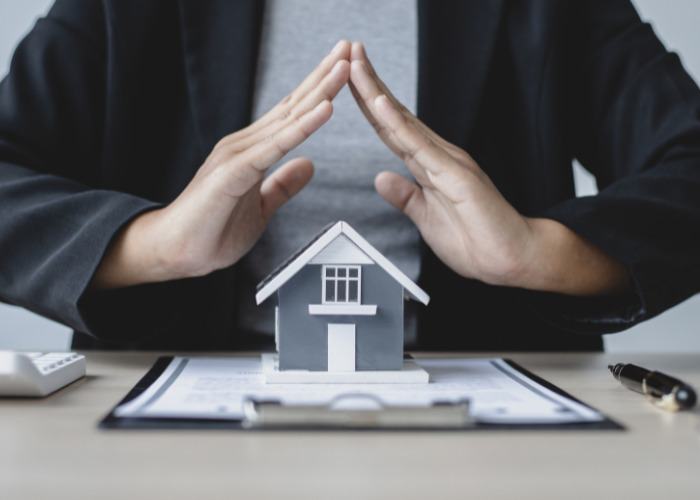Dłonie tworzą dach nad modelem domu na umowie, symbolizując ochronę. W tle kalkulator i długopis, sugerujące kalkulacje i formalności związane z ubezpieczeniem.