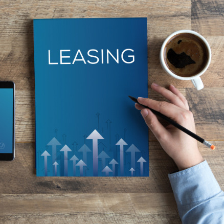 Niebieska teczka z napisem LEASING i strzałkami w górę, telefon, kawa i dłoń z ołówkiem na drewnianym biurku. Widok z góry, biznesowy minimalizm.