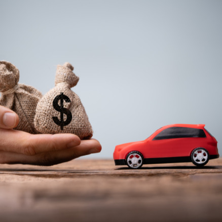 Dłoń trzyma dwa worki z symbolem dolara obok czerwonego modelu auta na drewnianym stole, sugerując finansowanie zakupu pojazdu.