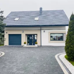 Nowoczesny dom jednorodzinny z grafitową dachówką, ciemnymi oknami, garażem i podjazdem z kostki brukowej, ozdobiony zielenią.