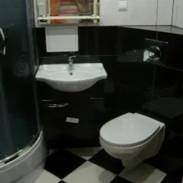 Nowoczesna łazienka z czarno-białą podłogą, kabiną prysznicową i wiszącą toaletą. Umywalka z szafką i czarne płytki na ścianach. Minimalistyczny design.