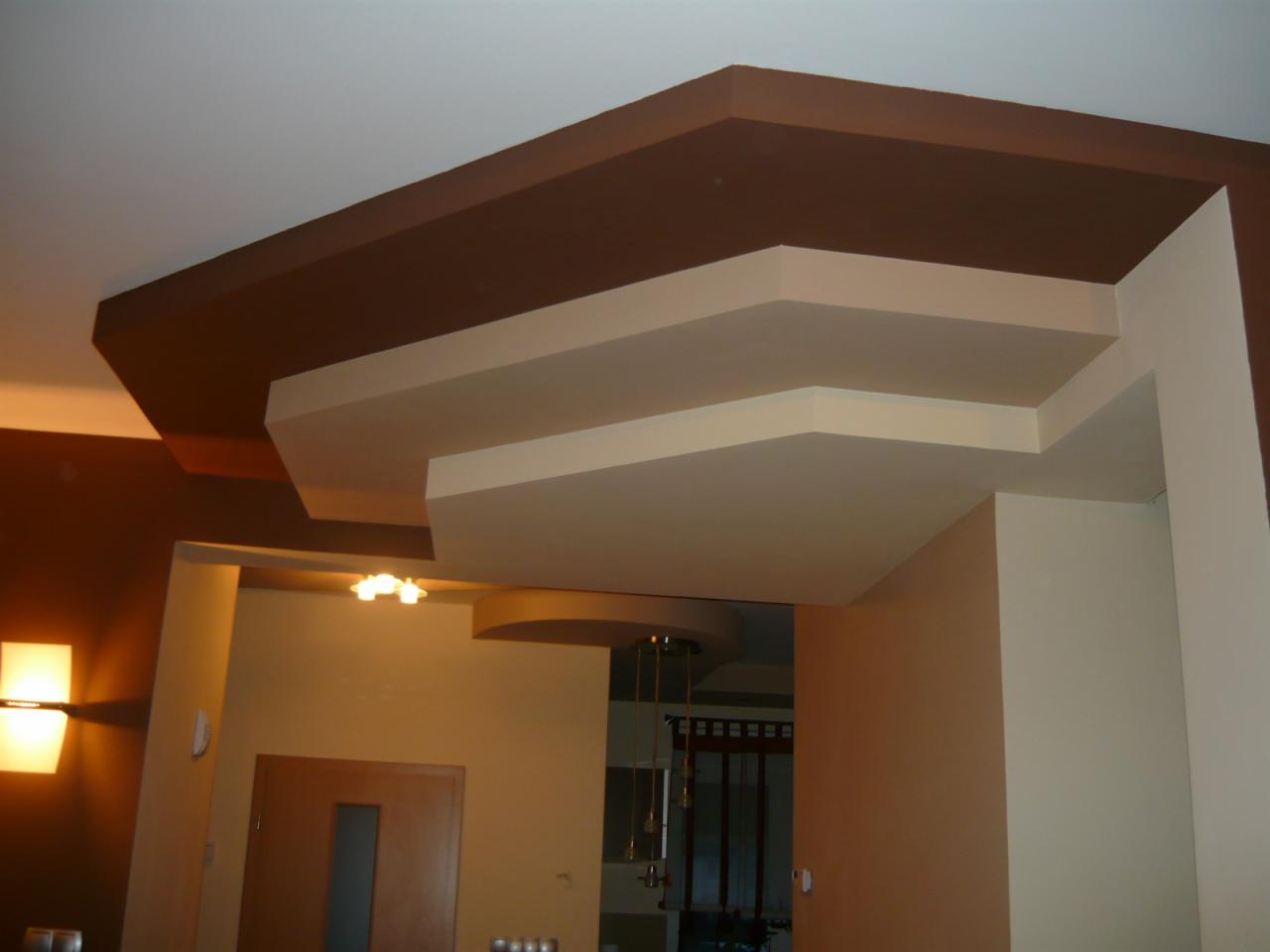 Wielopoziomowy sufit podwieszany z płyt kartonowo-gipsowych, pomalowany na beżowo i brązowo, z widocznym oświetleniem w tle. Dekoracyjne wykończenie wnętrza.