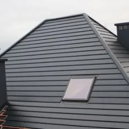 Szary dach z blachy na rąbek stojący z oknem dachowym i dwoma kominami, widok z dołu na tle pochmurnego nieba.
