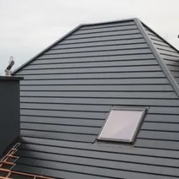 Fragment dachu z szarej blachy z oknem dachowym i kominami, widoczna konstrukcja więźby dachowej w trakcie prac.