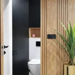 Nowoczesna toaleta z czarną ścianą, drewnianą okładziną pionową i rośliną doniczkową w złotym stojaku, widoczna z korytarza przez otwarte drzwi.