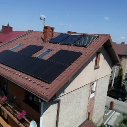 Instalacja paneli słonecznych i kolektorów słonecznych na brązowym dachu domu jednorodzinnego, widok z góry na tle błękitnego nieba.