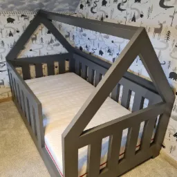 Szare łóżko dziecięce w kształcie domku z materacem, na tle ściany w leśne wzory.