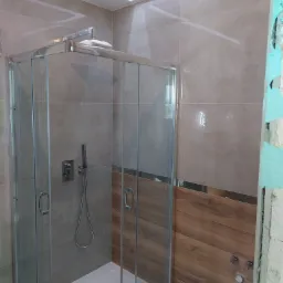 Kabina prysznicowa z chromowanymi elementami i szklanymi drzwiami, widoczne szare płytki oraz fragment drewnopodobnej okładziny, ściana w trakcie wykończenia z widocznym zabezpieczeniem folią.