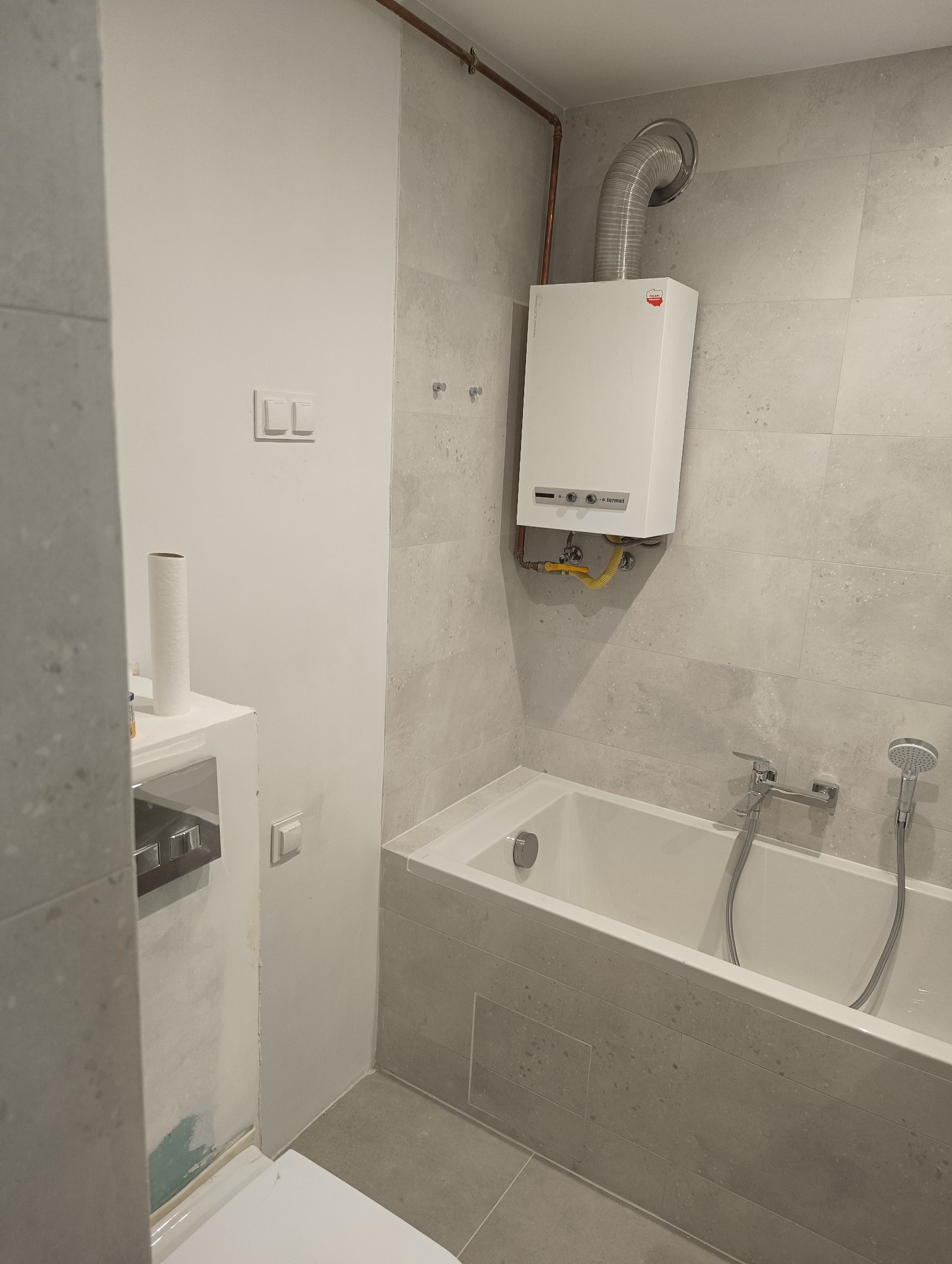 Szarym kamieniem wyłożona łazienka z wanną, baterią i prysznicem, piec gazowy z rurą odprowadzającą spaliny, widoczne przyłącza gazowe.