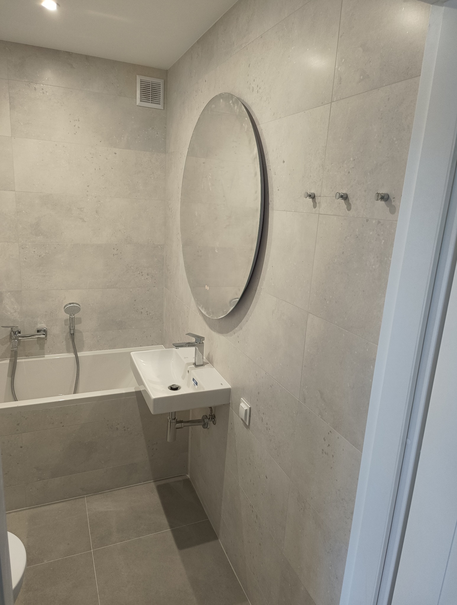 Jasna łazienka z owalnym lustrem, wiszącą umywalką i wanną, wykończona płytkami w odcieniach szarości z delikatną fakturą.