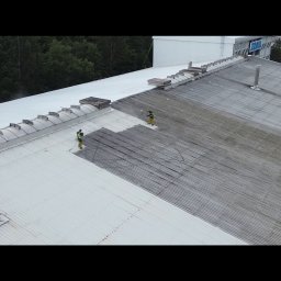 Malowanie dachów, Artdachmal - Dwóch pracowników w kombinezonach czyści dach hali przemysłowej za pomocą myjek ciśnieniowych. Widoczny fragment dachu przed i po czyszczeniu.