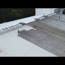 Dwóch pracowników w kombinezonach czyści dach hali przemysłowej za pomocą myjek ciśnieniowych. Widoczny fragment dachu przed i po czyszczeniu.