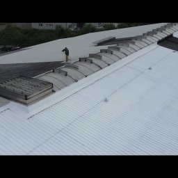 Malowanie dachów, Artdachmal - Malowanie dachu przemysłowego z widocznymi świetlikami. Dwóch pracowników w kombinezonach maluje dach na biało, widoczny kontrast między pomalowaną a niepomalowaną częścią dachu.